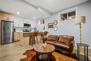 A modern kitchen with a refrigerator, microwave, and island with bar stools. There is a leather sofa with a coffee table and a cowhide rug. - `Stampede` King Bed | 7 Min to Downtown + Parking (Calgary)