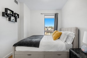 Ein Schlafzimmer mit einem Bett, einem Nachttisch, einer Lampe, einem Fenster mit Blick auf Gebäude und einem an der Wand befestigten Regal mit Pflanzen.