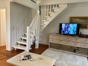 Ein Wohnzimmer mit weißer Treppe, einem Fernseher auf einer Holzkommode und einer Zeitschrift auf einem Couchtisch.