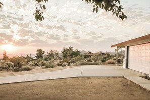 A residential driveway with a garage, surrounded by desert vegetation and houses in the background. - Hot tub I Fire pit I 10 min to park I 5 min to JT (Joshua Tree)