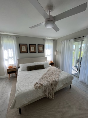 Une chambre à coucher avec un lit, un ventilateur de plafond, des tableaux encadrés accrochés au mur et une vue sur l’extérieur grâce à une porte vitrée.