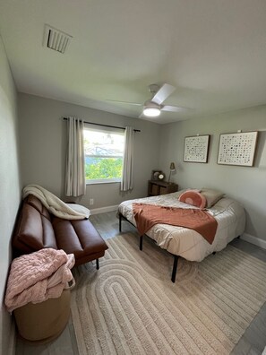 A bedroom with a bed, a couch, a nightstand, a ceiling fan, and a window with curtains. - Cozy 3-bedroom home near Warm Mineral Springs, mayakka river, and gulf beaches (North Port)