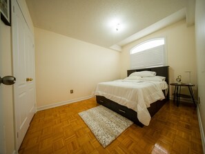 A bedroom with a bed, a small table, a clock, and a window with blinds. - 3-bedroom DUPLEX apartment in a serene environment Brampton, Canada (Brampton)