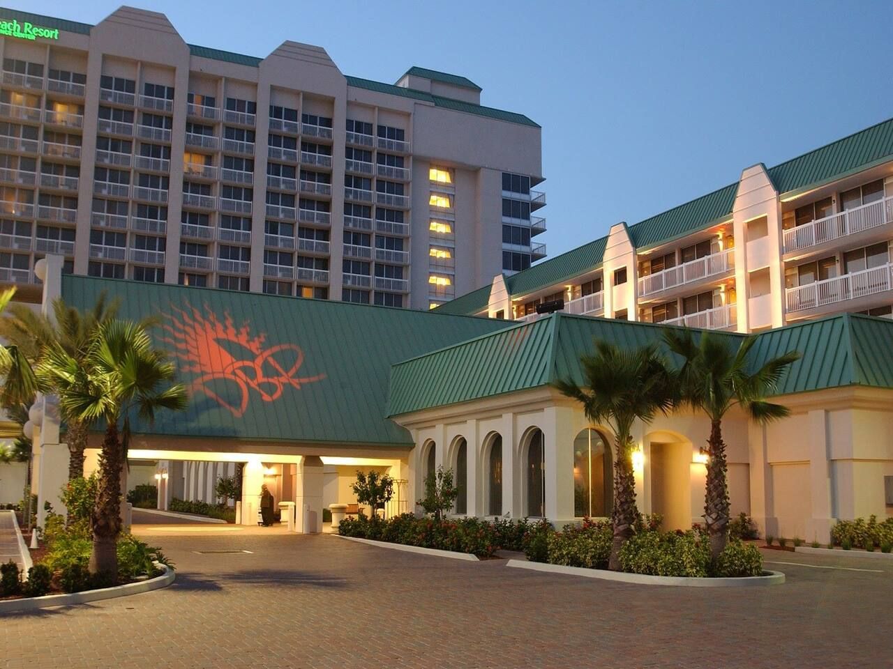 A hotel with a green roof, multiple balconies, and a palm tree-lined entrance.