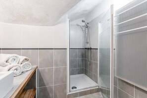 A modern bathroom with a glass-enclosed shower, grey tiles, and neatly rolled white towels on a wooden shelf. - Holiday Home 'Mas Bousquette' with Private Pool, Private Terrace and Wi-Fi (Lussan)
