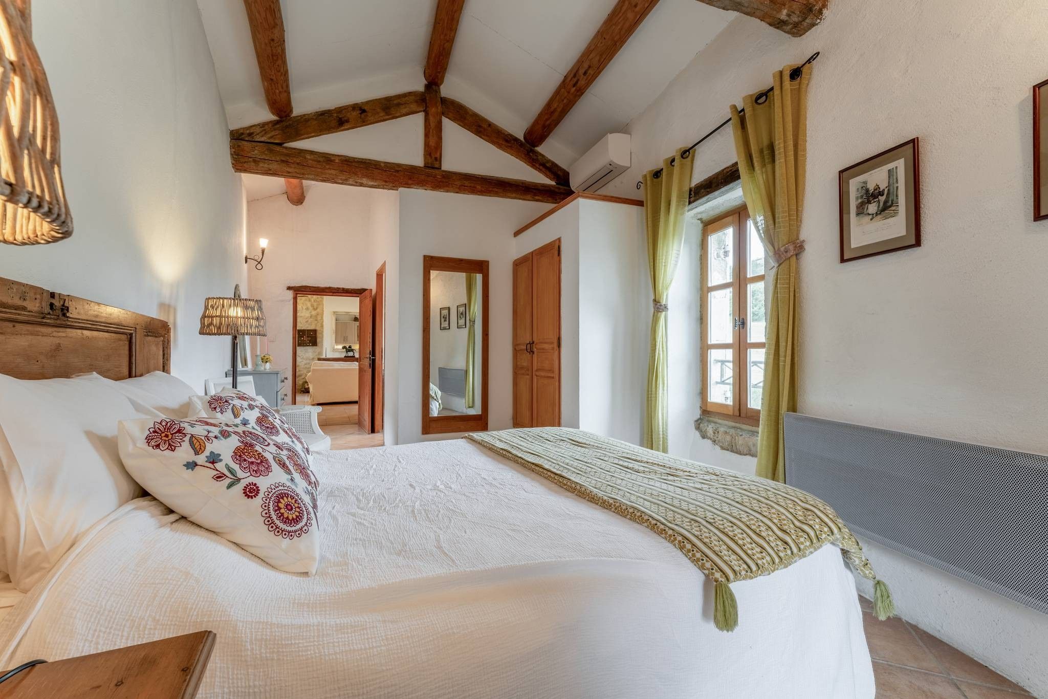 A bedroom with a large bed, wooden beams, a window with curtains, and a mirror.