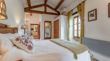 A bedroom with a large bed, wooden beams, a window with curtains, and a mirror.