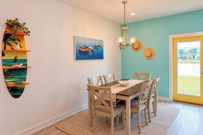 A dining area with a wooden table and chairs, a hanging light fixture, and a wall-mounted shelf with decorative items. - 3BR Townhome / Game Room & Pond Views / 5-Min Drive to Beach / PCB (Panama City Beach)