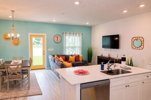 A modern kitchen with a built-in dishwasher, a dining area with a table and chairs, a sofa with cushions, a clock on the wall, and a TV mounted above the sink. - 3BR Townhome / Game Room & Pond Views / 5-Min Drive to Beach / PCB (Panama City Beach)