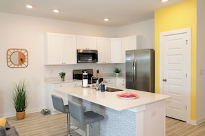 A modern kitchen with white cabinets, stainless steel appliances, a central island with bar stools, and a yellow accent wall. - 3BR Townhome / Game Room & Pond Views / 5-Min Drive to Beach / PCB (Panama City Beach)