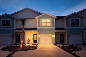 A two-story residential building with a central entrance, garage doors, and a landscaped front yard. - 3BR Townhome / Game Room & Pond Views / 5-Min Drive to Beach / PCB (Panama City Beach)