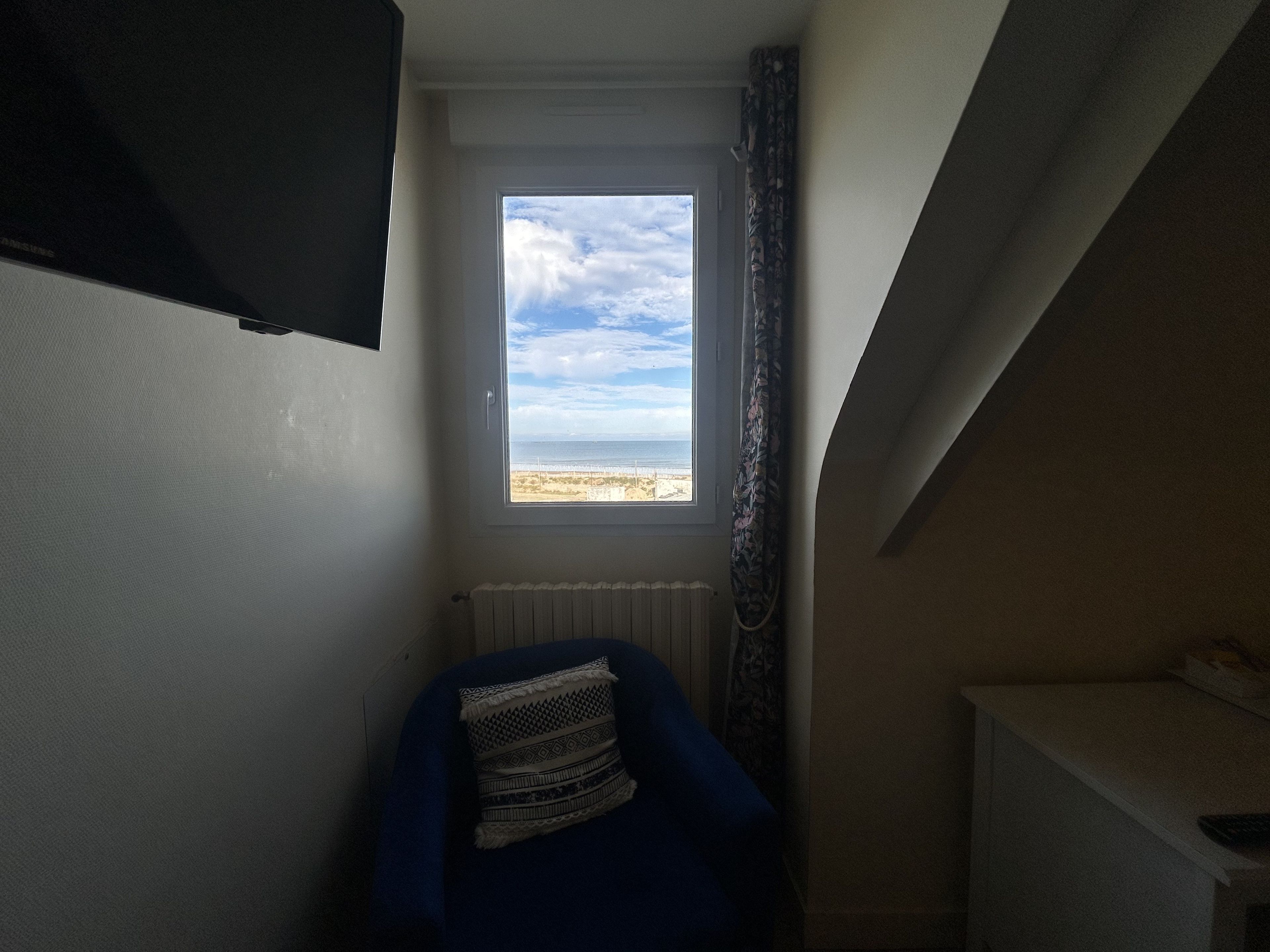A room with a window overlooking a beach, a radiator, and a blue chair with a patterned cushion.