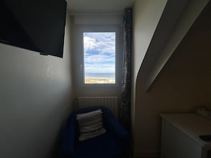 Habitació amb finestra que dona a una platja, un radiador i una cadira blava amb coixí estampat.
