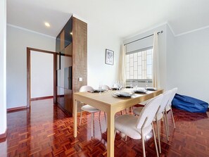 A dining area with a wooden table set for four, a sofa, and a large window with curtains. - Palmeira Apartment by Atlantic Holiday (Santa Cruz)