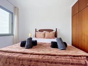 A neatly made bed with a wooden headboard, a window with curtains, and a wardrobe. - Palmeira Apartment by Atlantic Holiday (Santa Cruz)