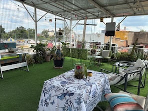 Een terras buiten met een tafel, overdekt met een tafelkleed met bloemenprint, omringd door stoelen en bloempotten.