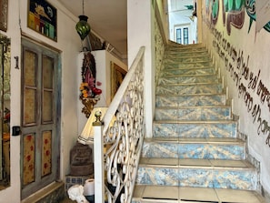 Un escalier orné de rampes décoratives et de carreaux aux motifs variés, menant à une pièce dotée d’une porte et d’un mur orné d’une fresque.