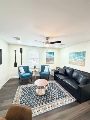 A living room with a black leather sofa, two blue armchairs, a round coffee table, and a patterned rug. There are two paintings on the wall, a ceiling fan, and a window with blinds. - Cozy Coastal 2 BDR Condo w/Parking Wildwood Crest (Wildwood Crest)