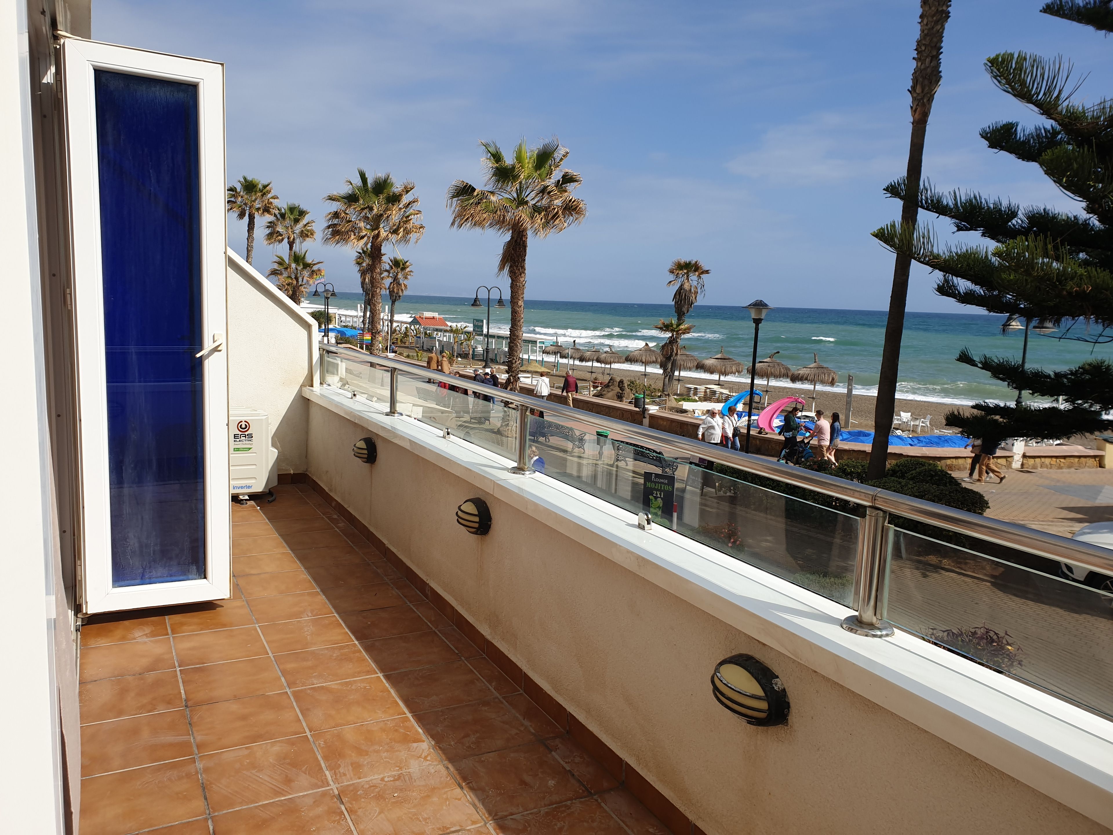 A balcony with a view of the beach, palm trees, and beach umbrellas.