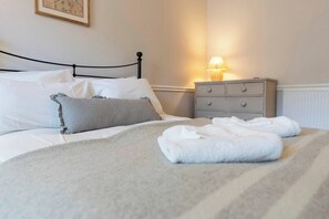 Ein ordentlich bezogenes Bett mit weißer Bettwäsche und einem gestreiften Kopfkissen, ein Nachttisch mit Lampe sowie eine Kommode mit Schubladen.