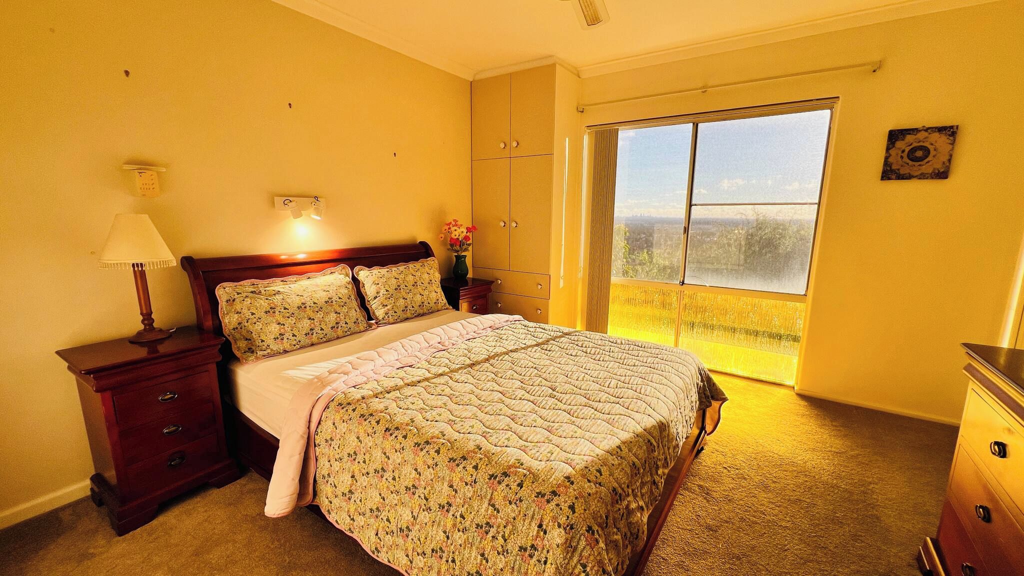 A bedroom with a bed, a nightstand, a dresser, a window with a view, and a wall-mounted light.