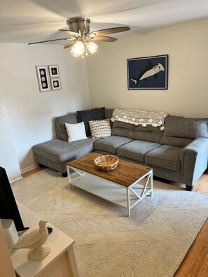 Ein Wohnzimmer mit Ecksofa, Couchtisch aus Holz, Deckenventilator und Wandbildern.