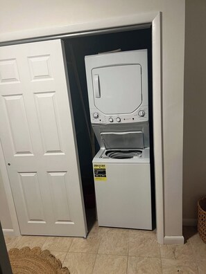 Un lavadero pequeño con lavadora y secadora apiladas, una puerta blanca y piso de baldosas.