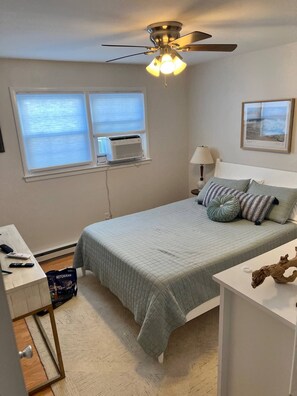 Un dormitorio con cama, escritorio, ventilador de techo, aire acondicionado, lámpara y un cuadro en la pared.