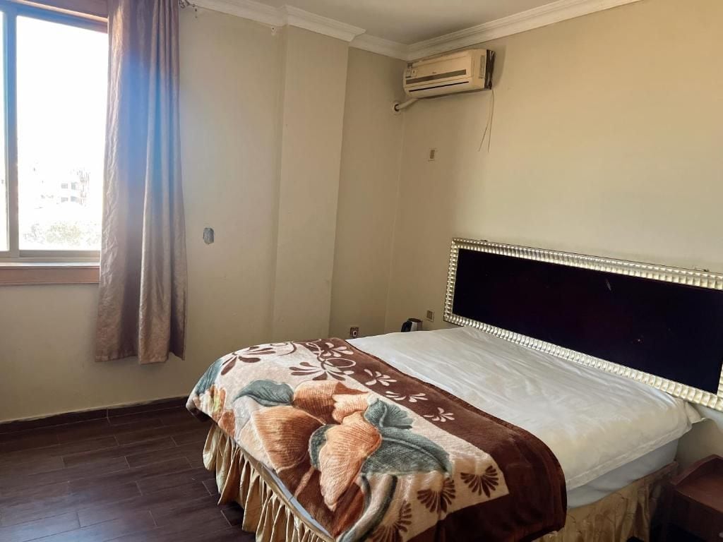 Un dormitorio con cama, unidad de aire acondicionado, ventana con cortinas y vista a edificios.