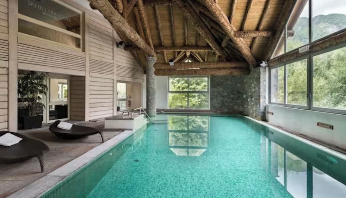 An indoor swimming pool with clear water, wooden ceiling beams, and a view of trees through large windows.