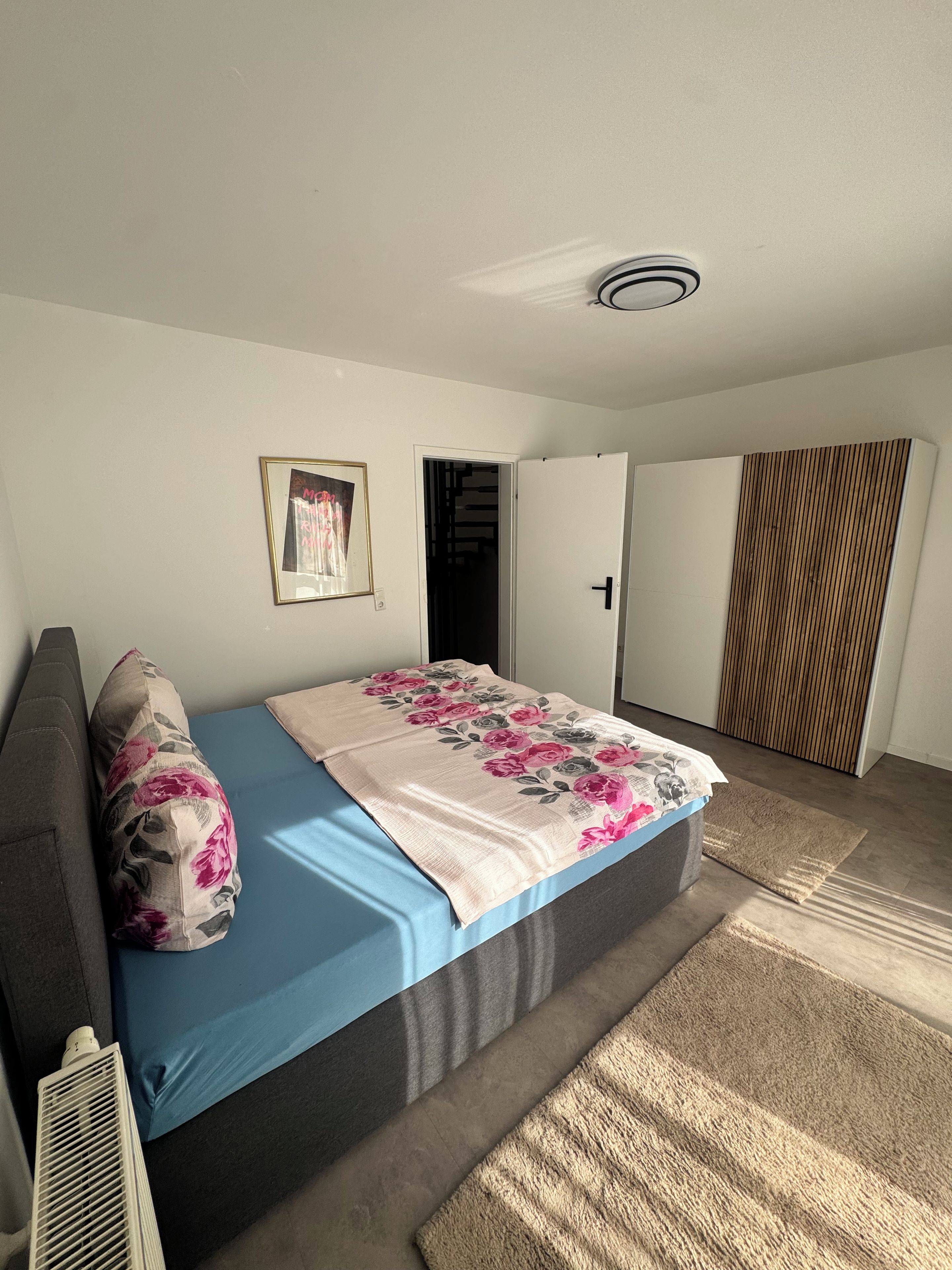 Une chambre à coucher avec un lit, une porte, un tableau accroché au mur et un luminaire fixé au plafond.