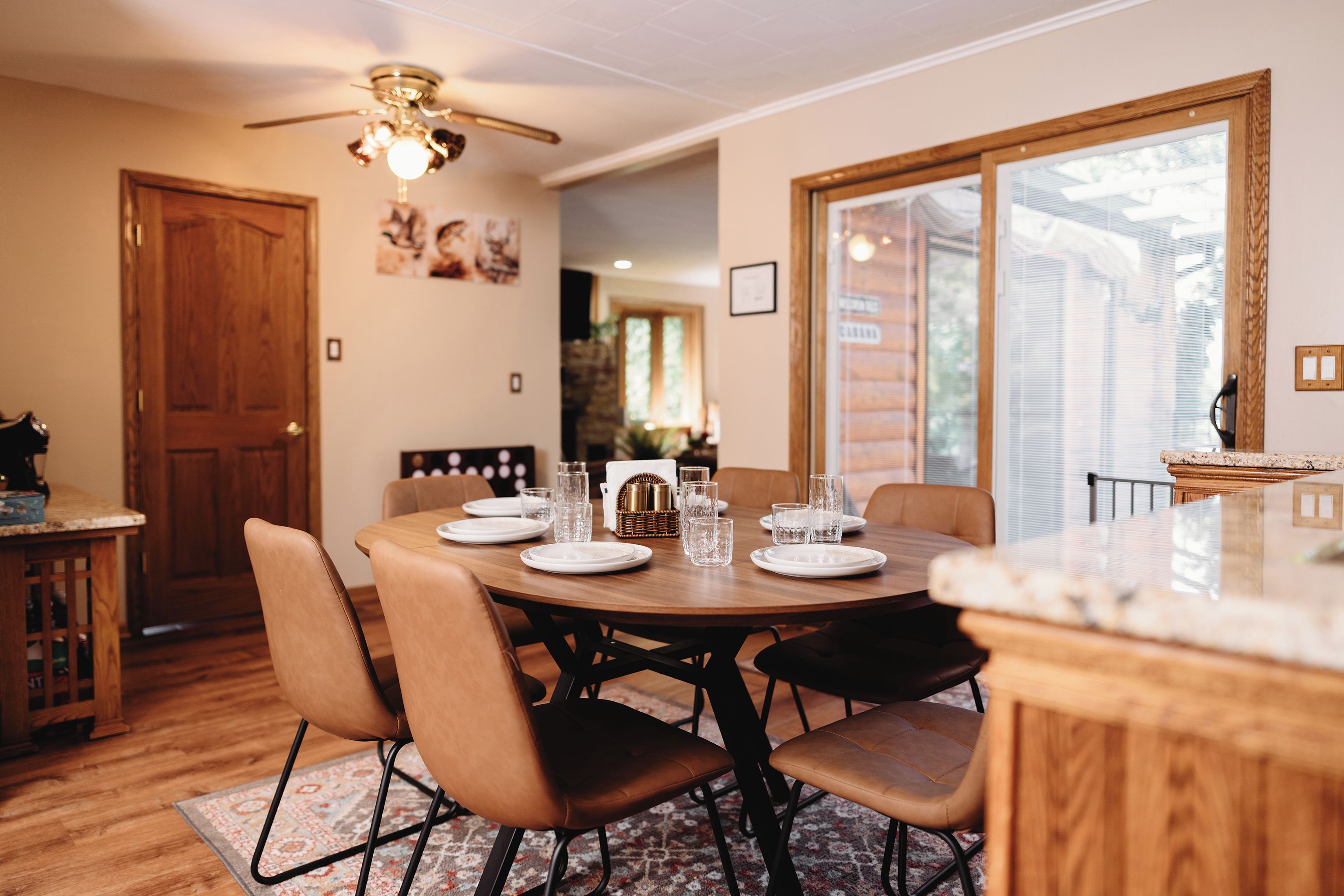 A dining area with a round table set for four, surrounded by chairs, with a ceiling fan above and a window to the right.