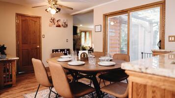 A dining area with a round table set for four, surrounded by chairs, with a ceiling fan above and a window to the right.