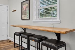 A room with a long wooden bench, three leather stools, a framed artwork, and a window with blinds. - Belmont Duplex | Shops, Dining & Entertainment (Charlotte)