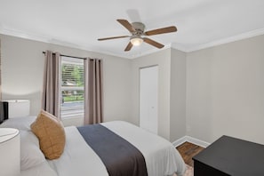 A bedroom with a bed, a ceiling fan, a window with curtains, a bedside table, and a door. - Belmont Duplex | Shops, Dining & Entertainment (Charlotte)