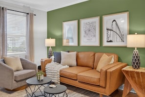 A living room with a brown leather sofa, a grey armchair, and a coffee table. There are three framed pictures on the wall and two lamps on either side of the sofa. - Belmont Duplex | Shops, Dining & Entertainment (Charlotte)