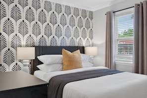 A hotel room with a bed, two bedside lamps, a window with curtains, and a patterned wallpaper. - Belmont Duplex | Shops, Dining & Entertainment (Charlotte)