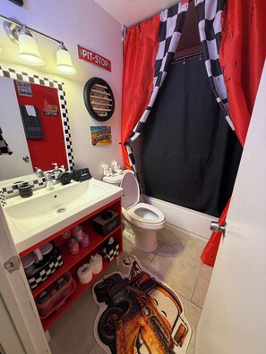 A bathroom with a red and black checkered shower curtain, a white toilet, a sink with a red shelf, and a mirror with a 'Pit Stop' sign. - Charming 2-bedroom apartment with AC in lively Phoenix (Phoenix)