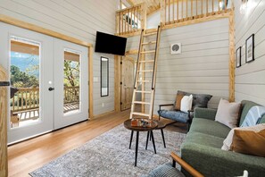 A living room with a wooden ladder, a television, a sofa, and a view through a door. - Green Getaway Cabin with Pasture Views (Chattanooga)