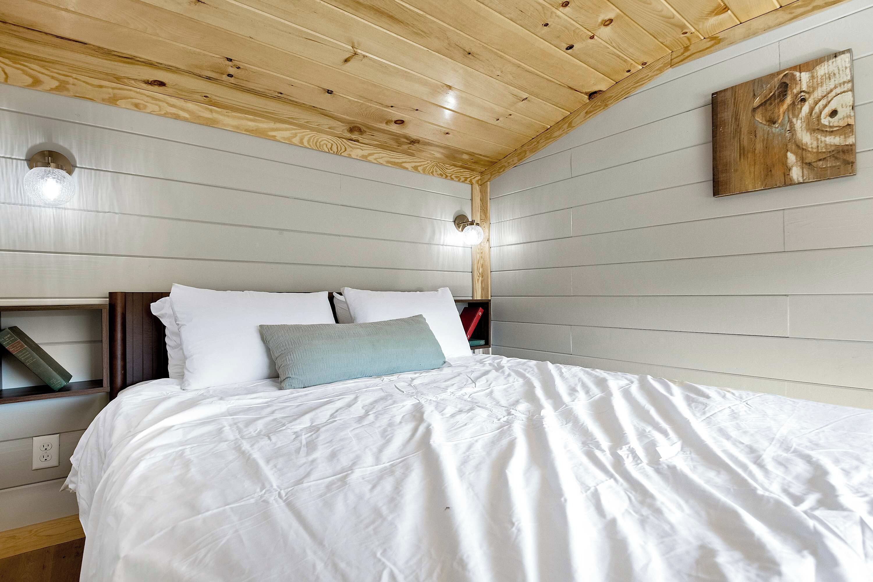 A bedroom with a wooden ceiling, a bed with white bedding, a nightstand with books, and a wall-mounted light fixture.