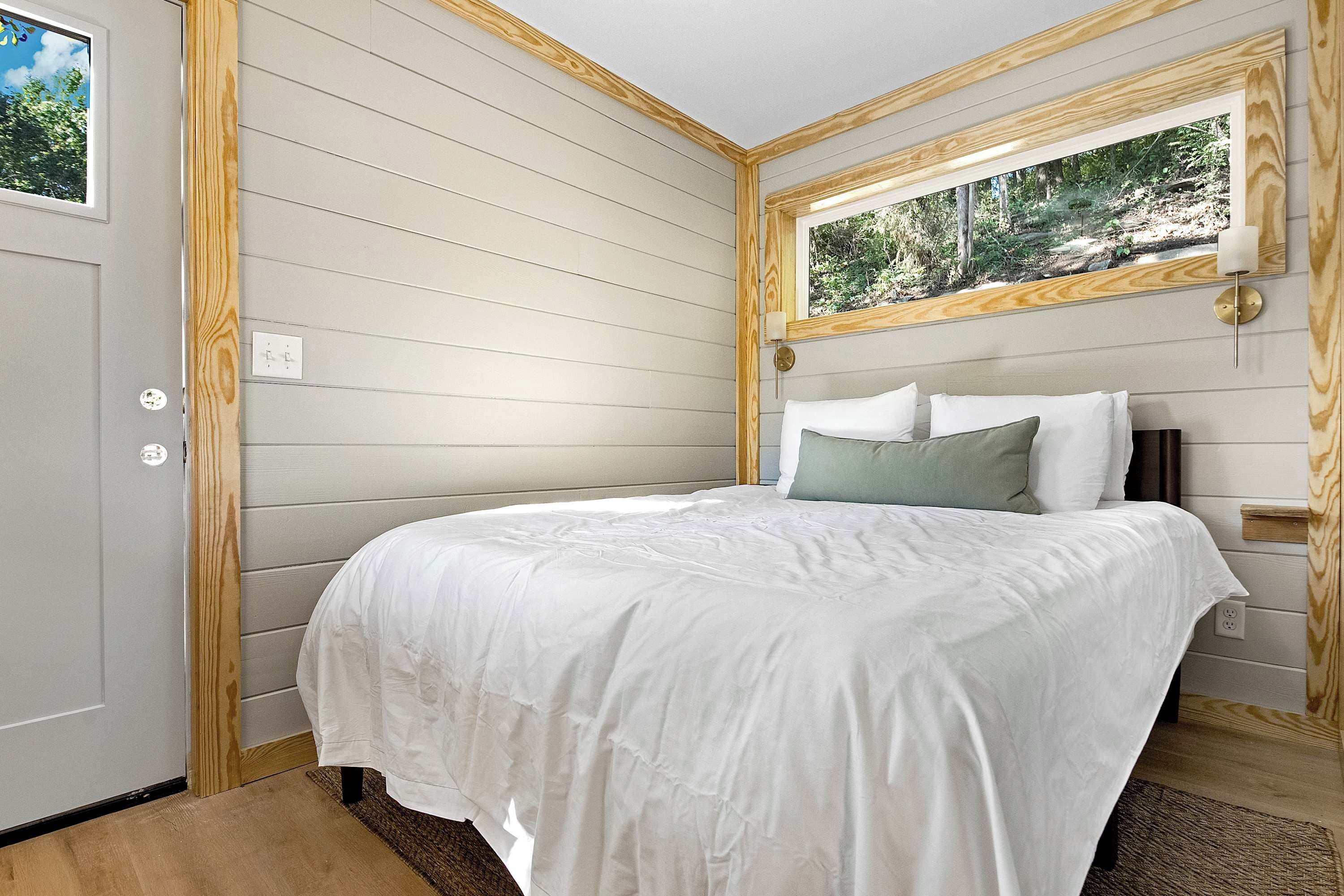 A bedroom with a bed, a window with a view of greenery, and a wall-mounted light fixture.