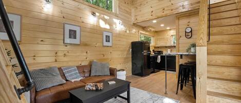 Interior de una cabaña de madera con sofá, una mesa, un refrigerador y zona de cocina.