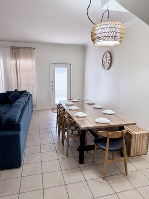 Uma sala de jantar com uma longa mesa de madeira posta para seis pessoas, com cadeiras, um sofá e um lustre.