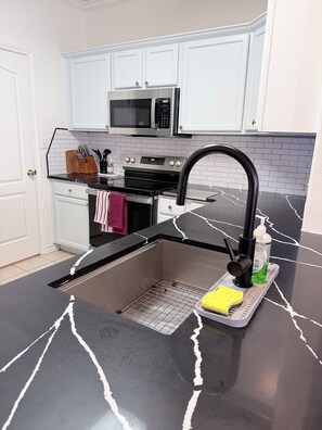 A kitchen with white cabinets, a stainless steel microwave, a black faucet, and a sink with a sponge and dish soap. - Full Renovated Coastal Retreat, Walk to Beach, Modern, Pool, Patio, Balcony. (Corpus Christi)