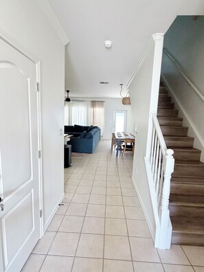 A hallway leading to a living room with a blue sofa, a dining area with a table and chairs, and a staircase with wooden steps. - Full Renovated Coastal Retreat, Walk to Beach, Modern, Pool, Patio, Balcony. (Corpus Christi)