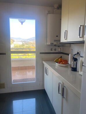 Une cuisine donnant sur un balcon et sur les montagnes, avec des meubles blancs et un saladier de fruits posé sur le plan de travail.
