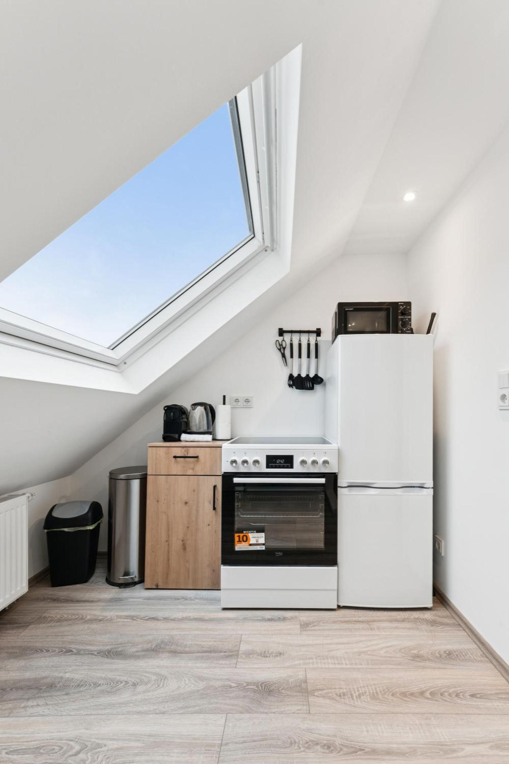 A compact kitchen with a skylight, a stove, a refrigerator, and a small trash bin.