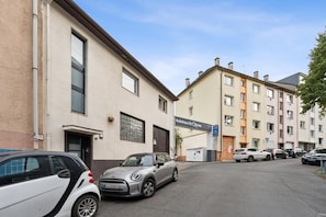 Straßenansicht mit einer Reihe mehrstöckiger Gebäude, einem vor der Tür stehenden Mini Cooper und einem Schild für 'Autohaus Ki Qhnen' auf der rechten Seite.