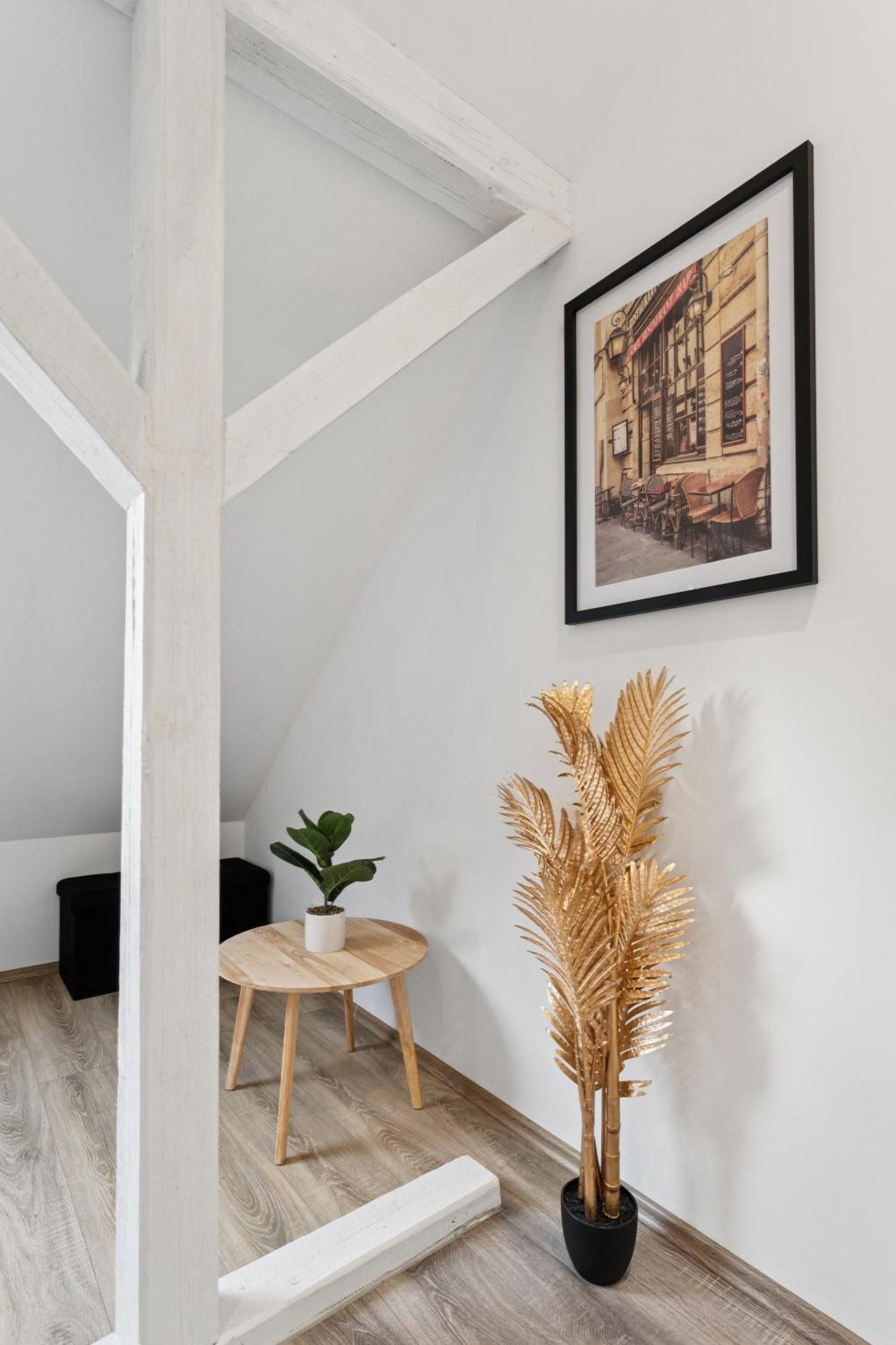 Eine Treppe mit weiß gestrichenen Balken, ein kleiner runder Holztisch mit einem Blumentopf und eine hohe, goldfarbene Zierpflanze im schwarzen Topf.