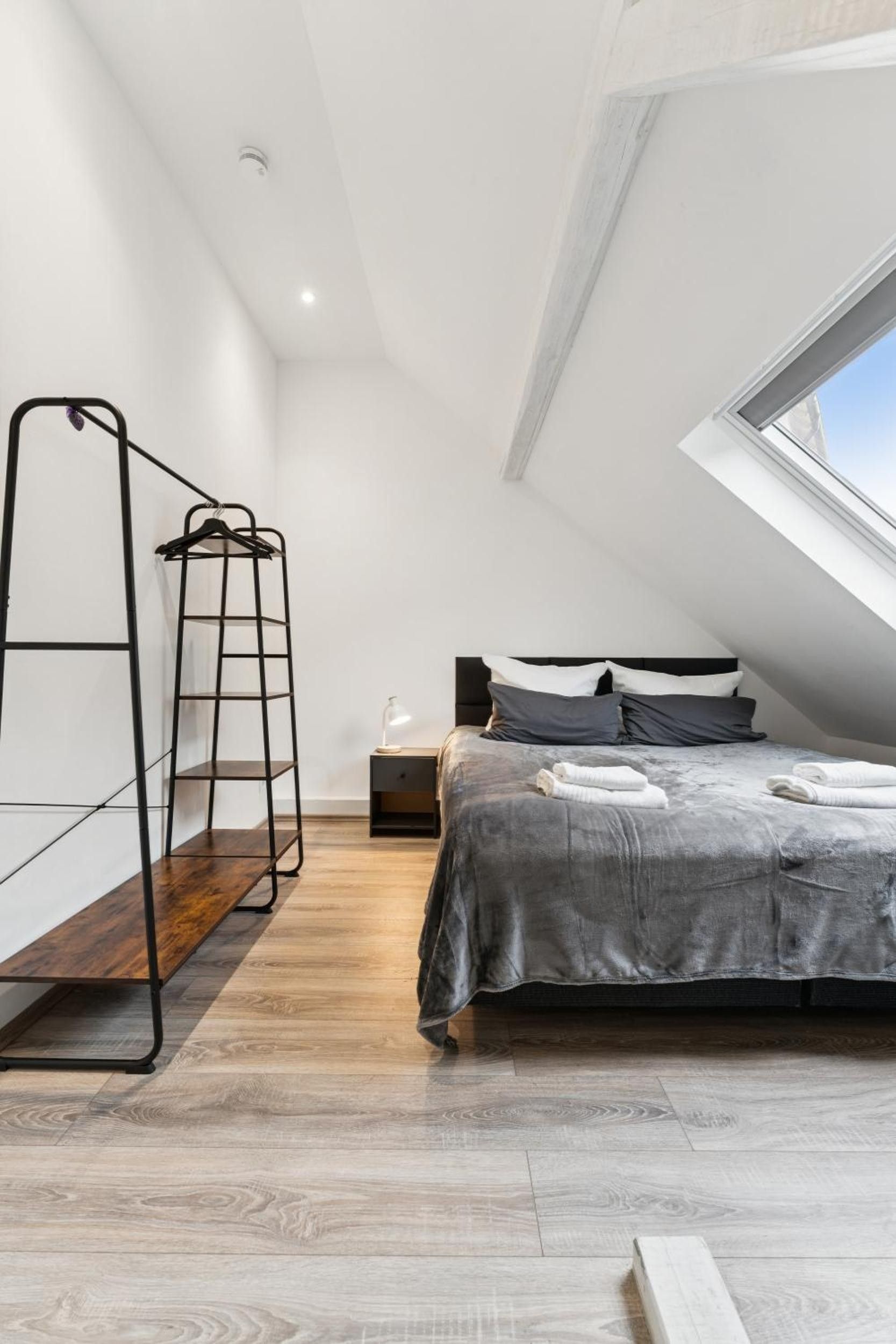 Ein modernes Schlafzimmer mit schrägem Dach, einem Bett mit grauer Bettwäsche, einer Holzbank und einem Metallregal.
